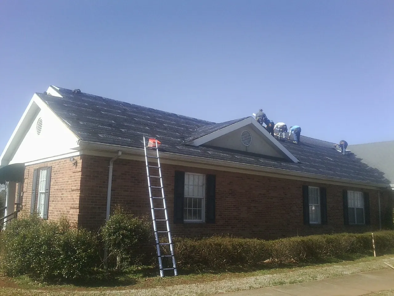 Crew tearing shingles off a brick ranch roof with a ladder set — Emergency Roof Repair work by our crew in Gregory