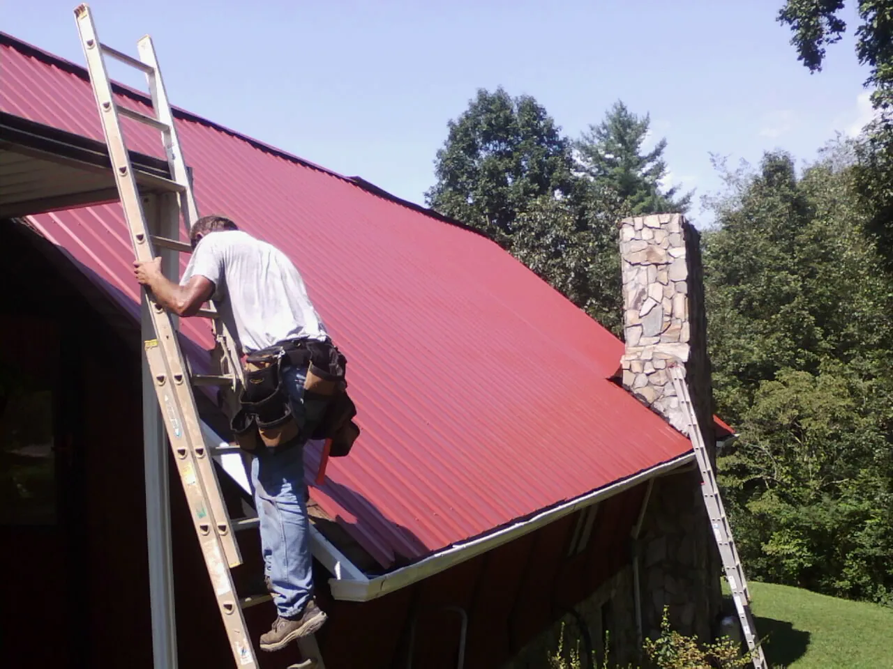 Roofer on a ladder beside a red metal roof and stone chimney — Soffit & Fascia Repair by Roofing Brothers in Gregory